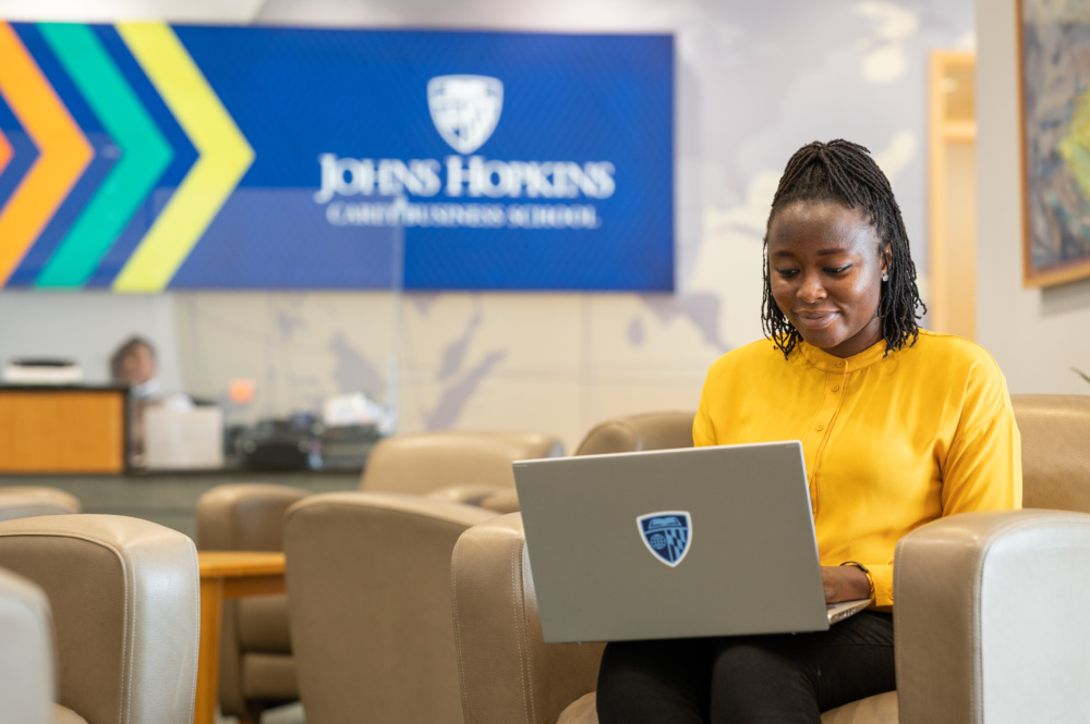 person sitting on couch with laptop, Carey Business School logo on tv in background