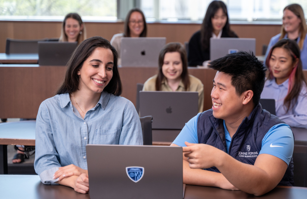 A group of students seated in a classroom, working on laptops. Other students are visible in the background, also on their laptops.