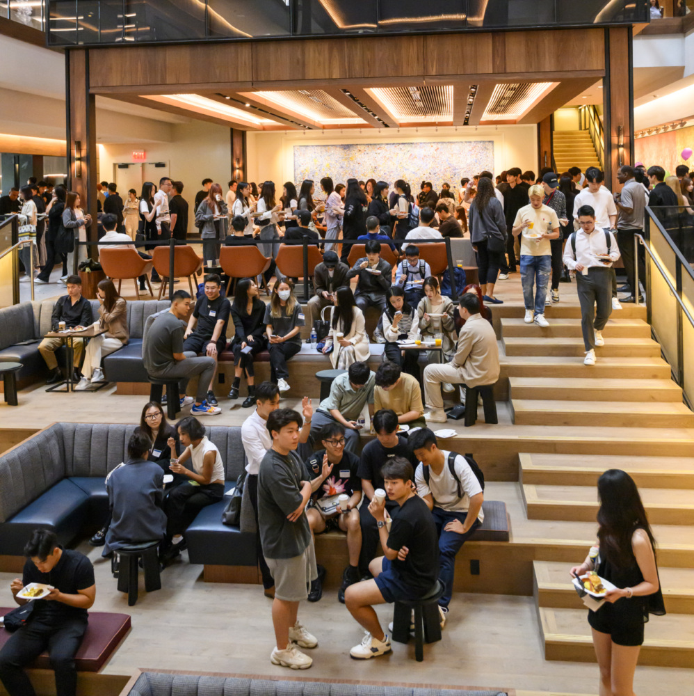 people standing and sitting on couches and at tables in a building with stairs and glass rooms