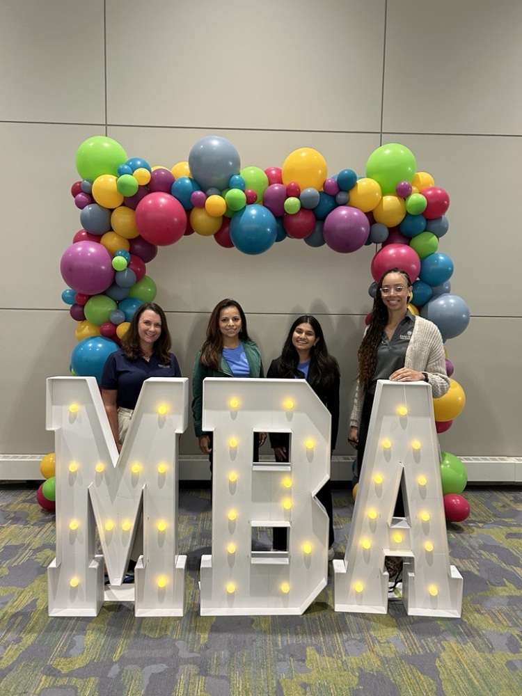 four people standing in back of big light up letters that spell &quot;MBA&quot; with a balloon arch in the back