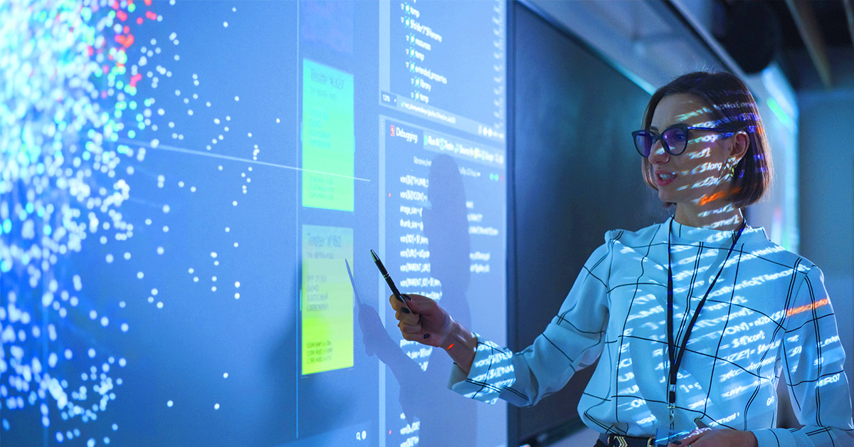 person standing in front of a whiteboard with a screen projector on the board and pointing to one of the graphs