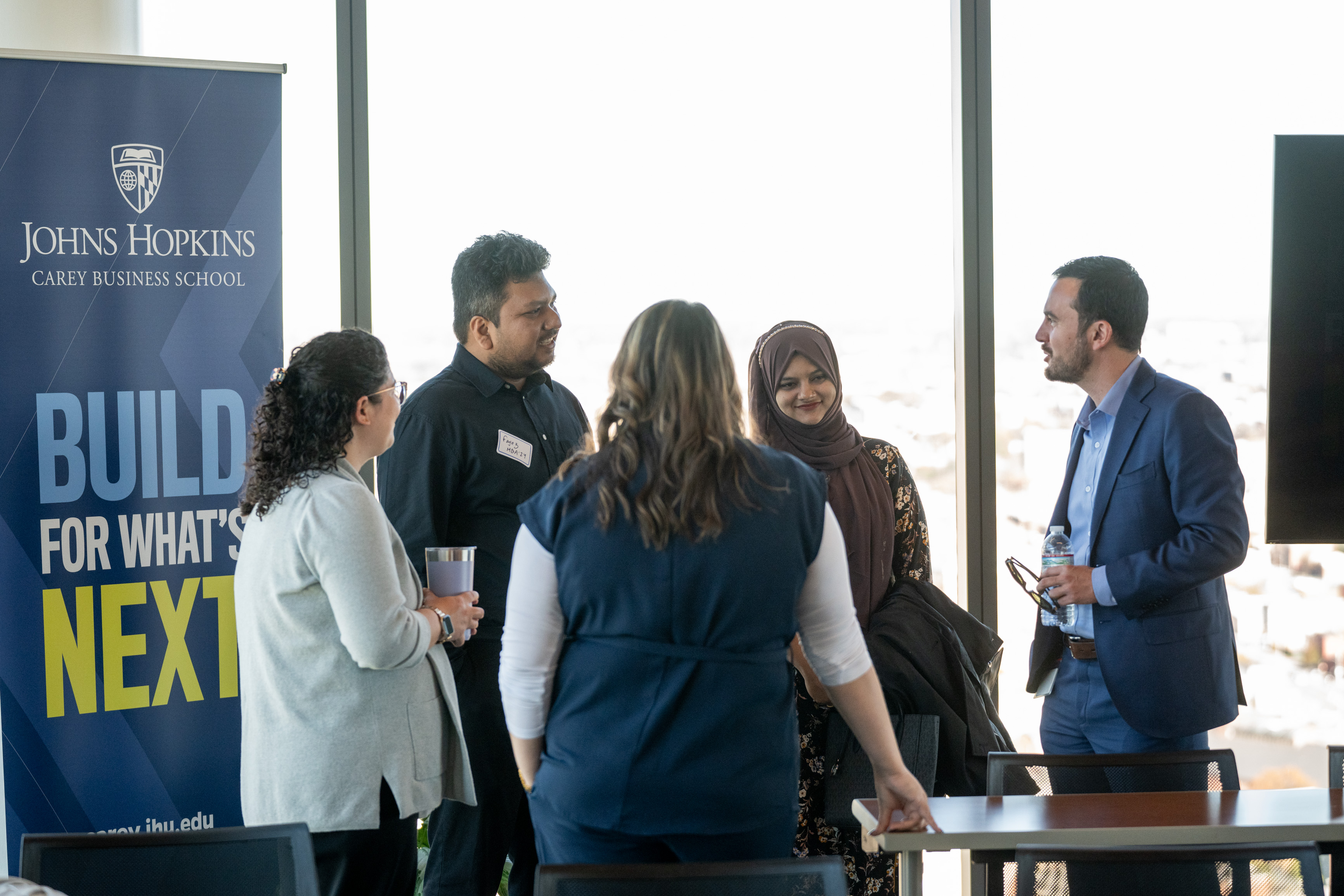 five people standing at a table speaking to each other. Johns Hopkins Carey Business School banner in the background