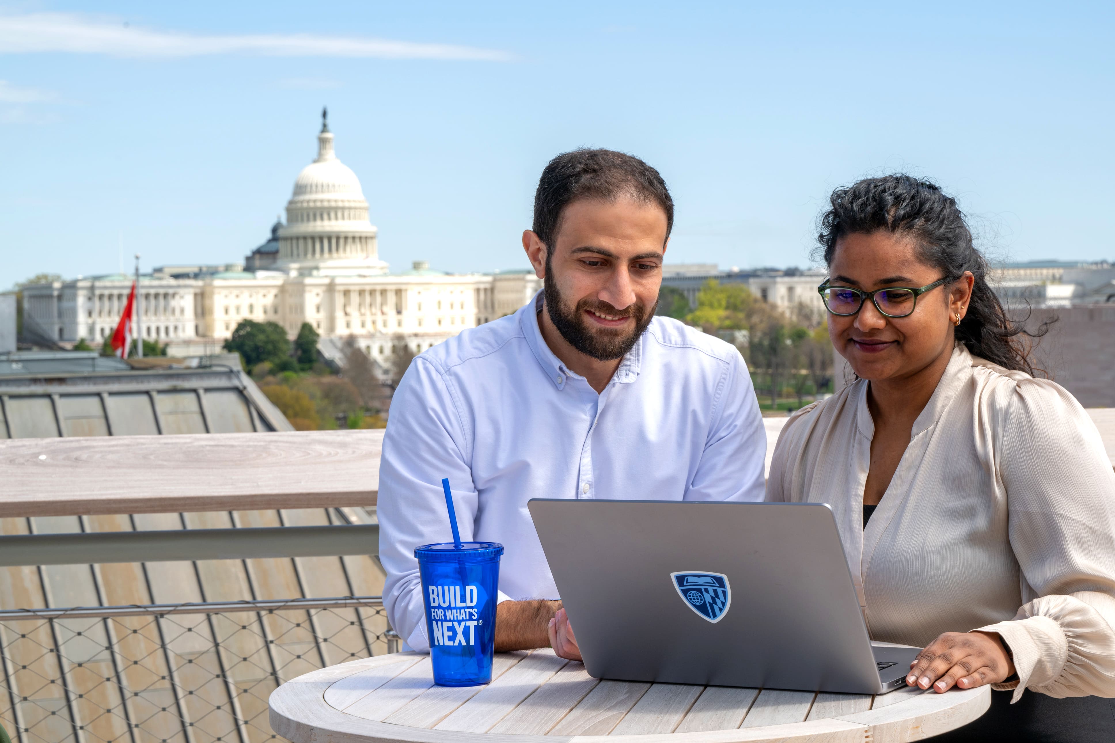 two people sitting outside at a table looking at a laptop outside with the capitol building in the background