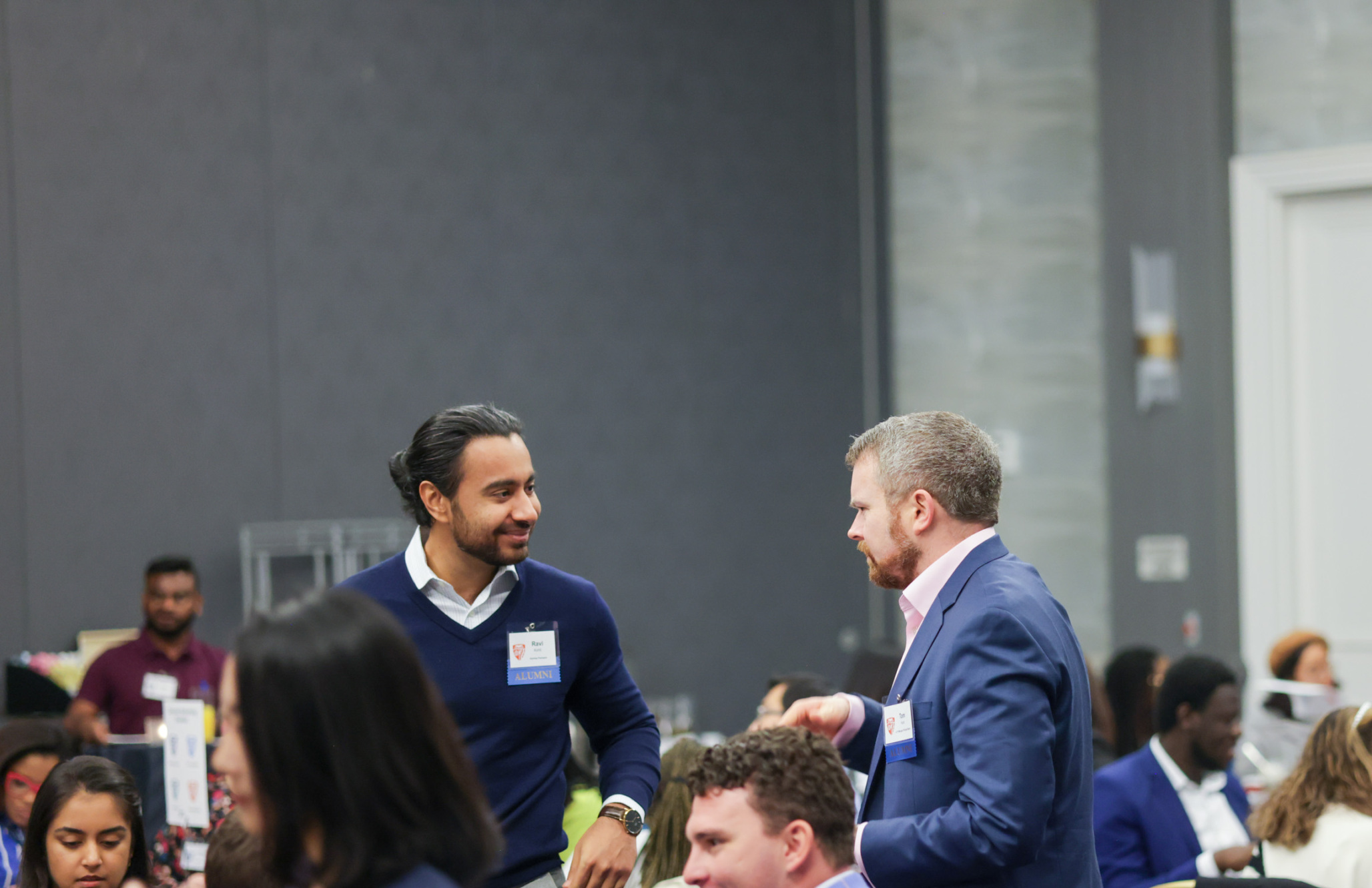 two people standing up in a group of people sitting at tables speaking to each other, smiling