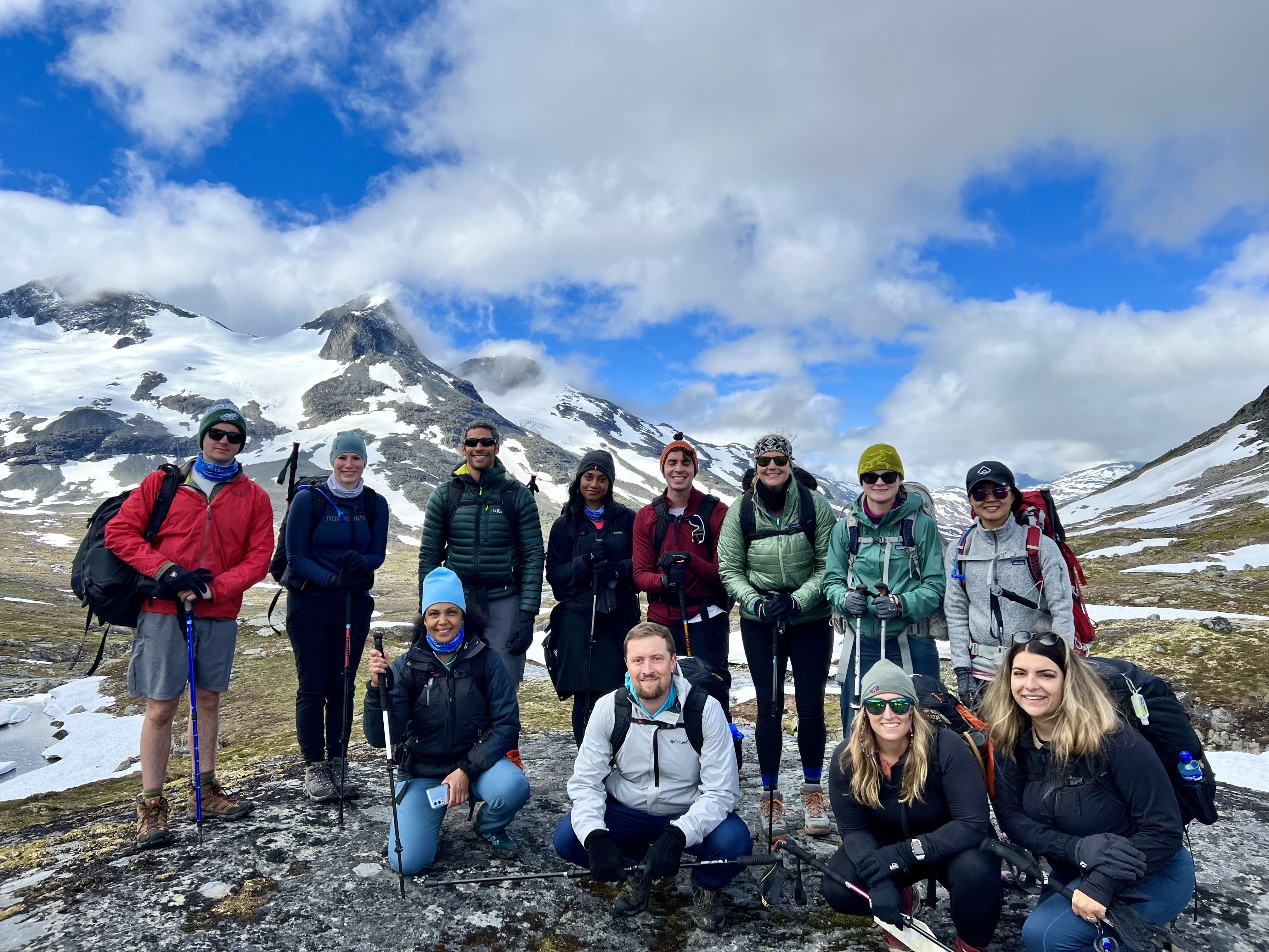 group of people posing for a picture outside on a mountain with snow and mountains in the background