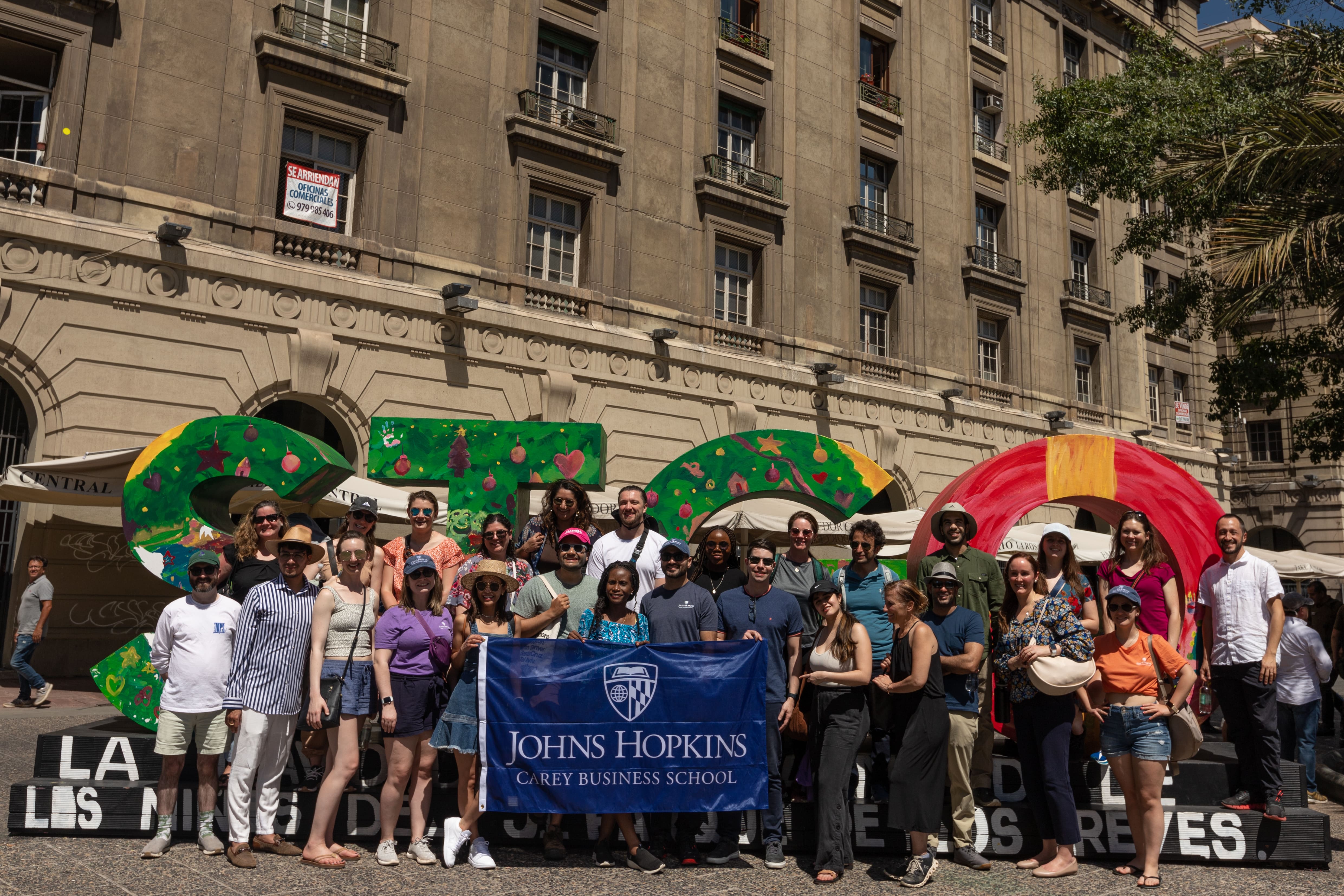 group of people standing outside in front of a building with a few of them holding a big flag that says Johns Hopkins Carey Business School