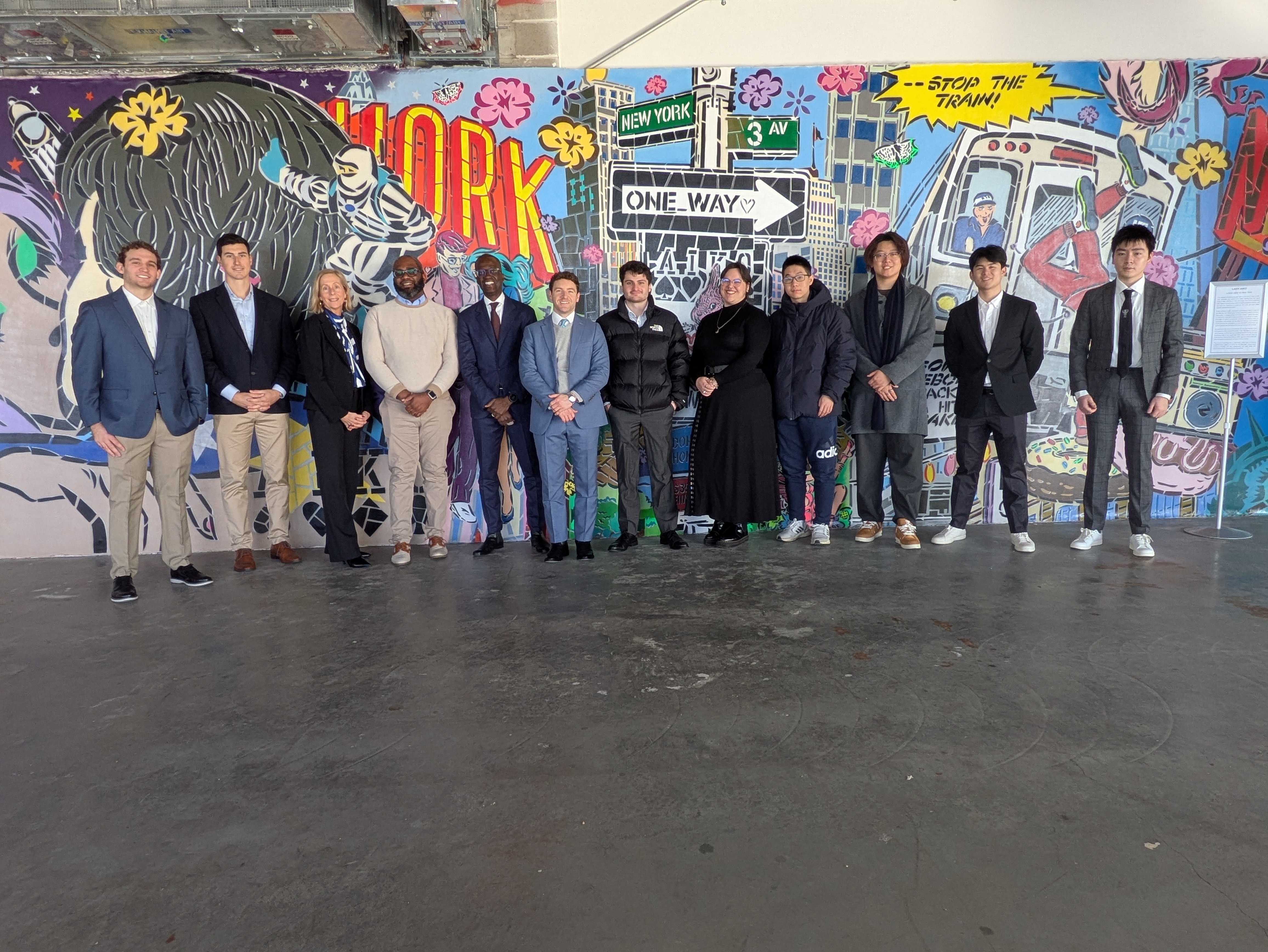 twelve people standing in a line posing for a photo. background is a new york painted mural