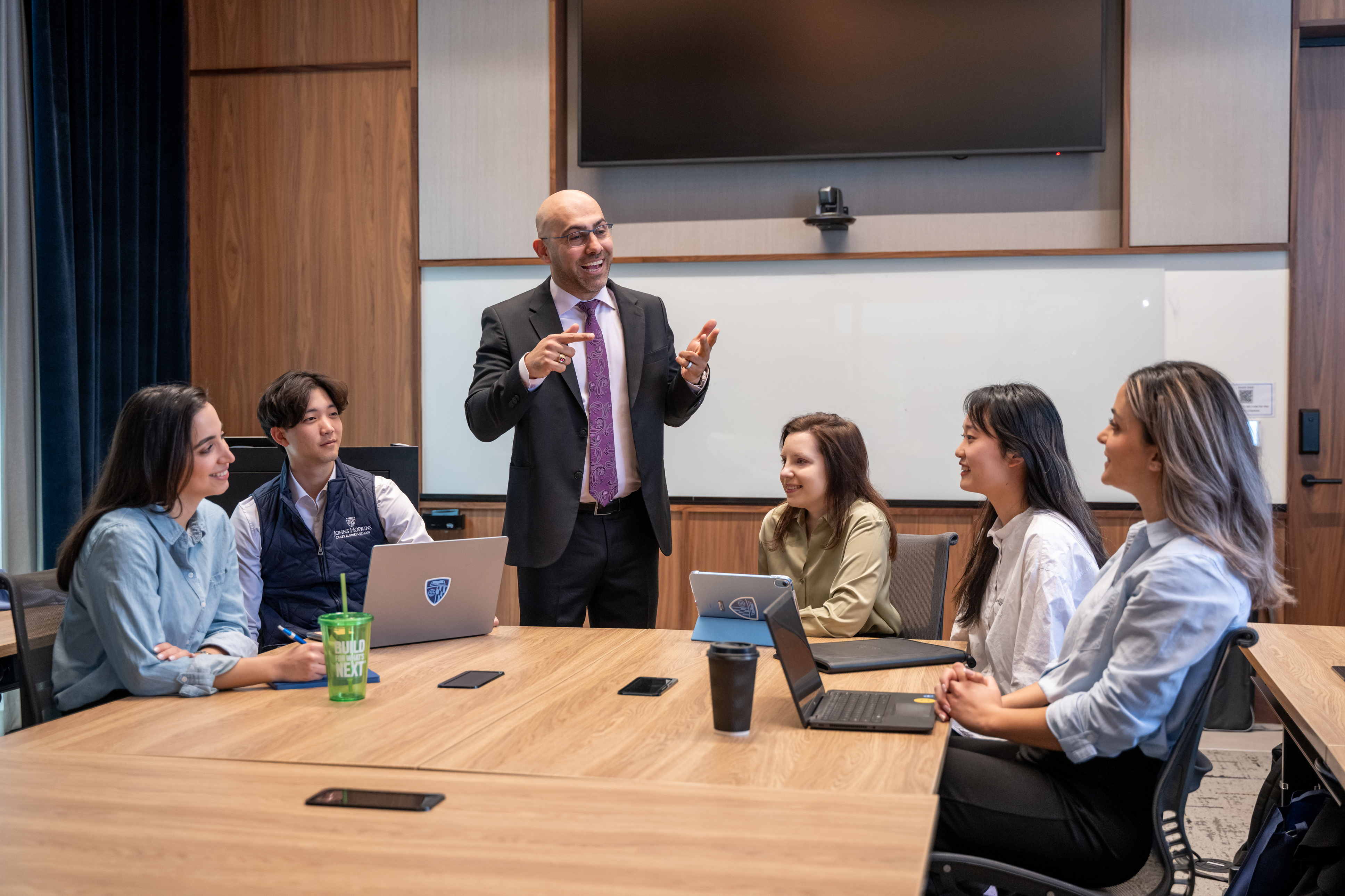 five people sitting at a table in a classroom and one person standing speaking