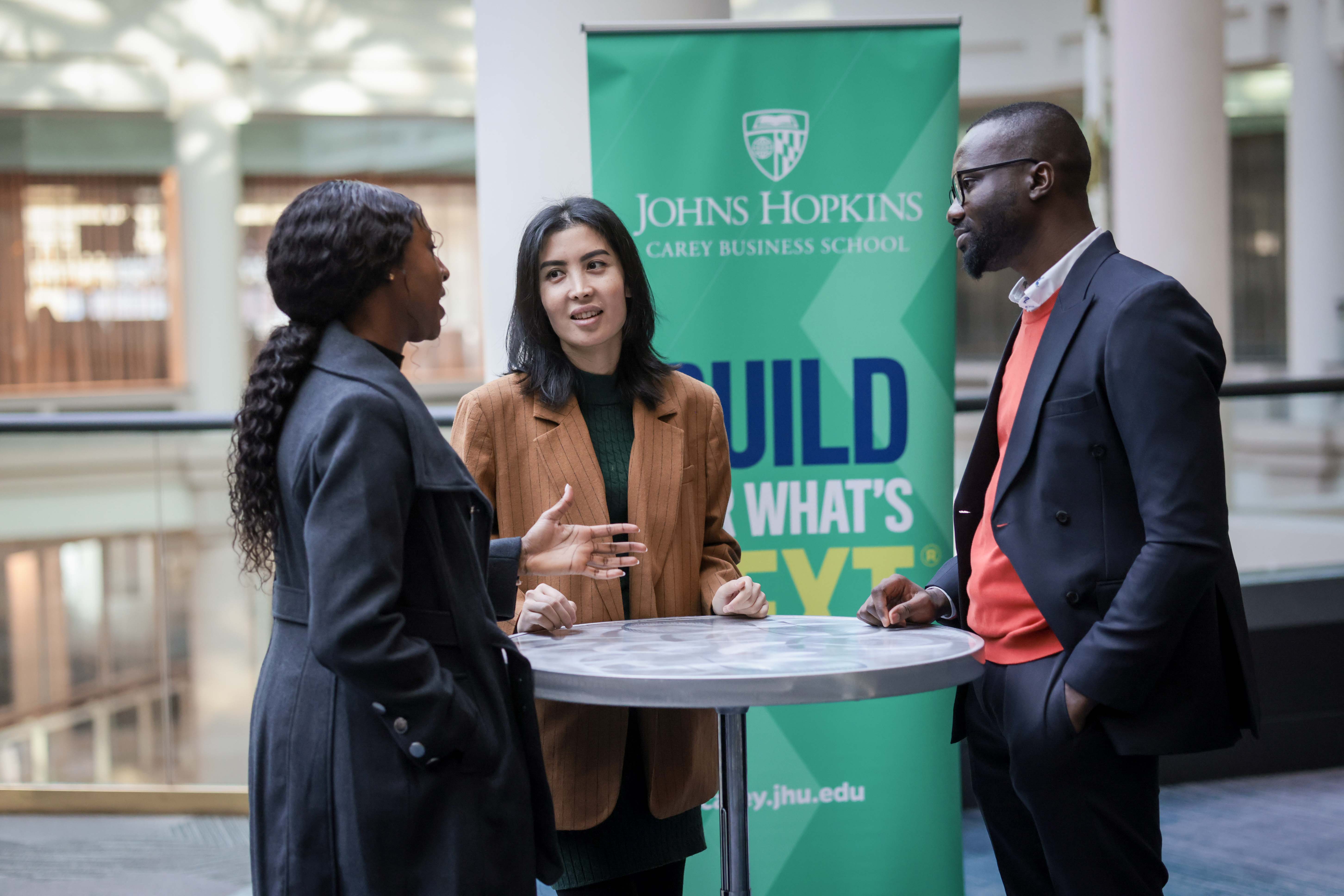 three people standing around a high top table talking with a johns hopkins carey business school banner in the background