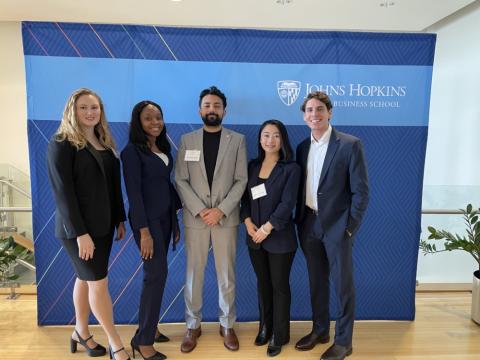 five students standing smiling for photo with Johns Hopkins banner in background