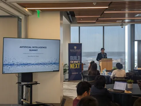 person standing at podium speaking into microphone with a room full of people sitting at tables