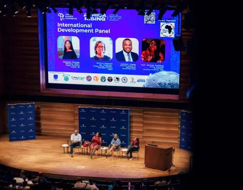 four people sitting in chairs on a stage. one person is holding and speaking into a microphone. a projector screen is in the background with their headshots