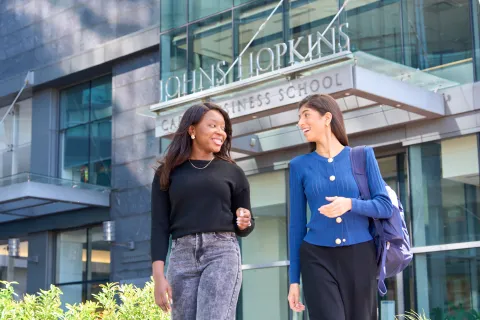 two people walking outside, looking at each other and talking. Johns Hopkins Carey Business School building in the back