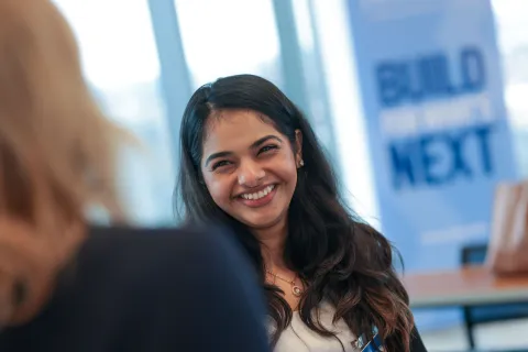 person smiling while talking to another person with a build for what's next banner in the back