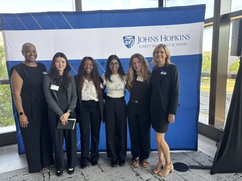 six people standing posing for a photo with the johns hopkins carey business school backdrop 