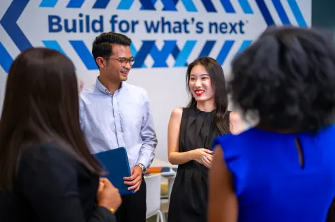 four people standing talking, 2 in view, other two view is of the back of their heads. smiling with a build for what's next sign in the background