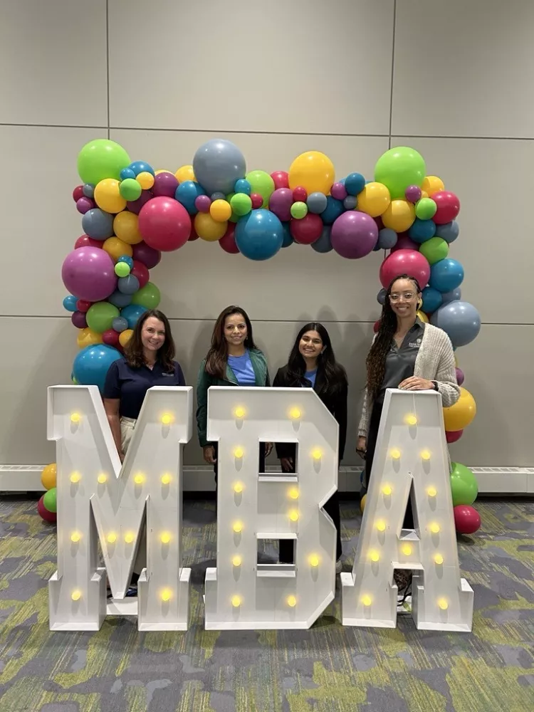 four people standing in back of big light up letters that spell "MBA" with a balloon arch in the back