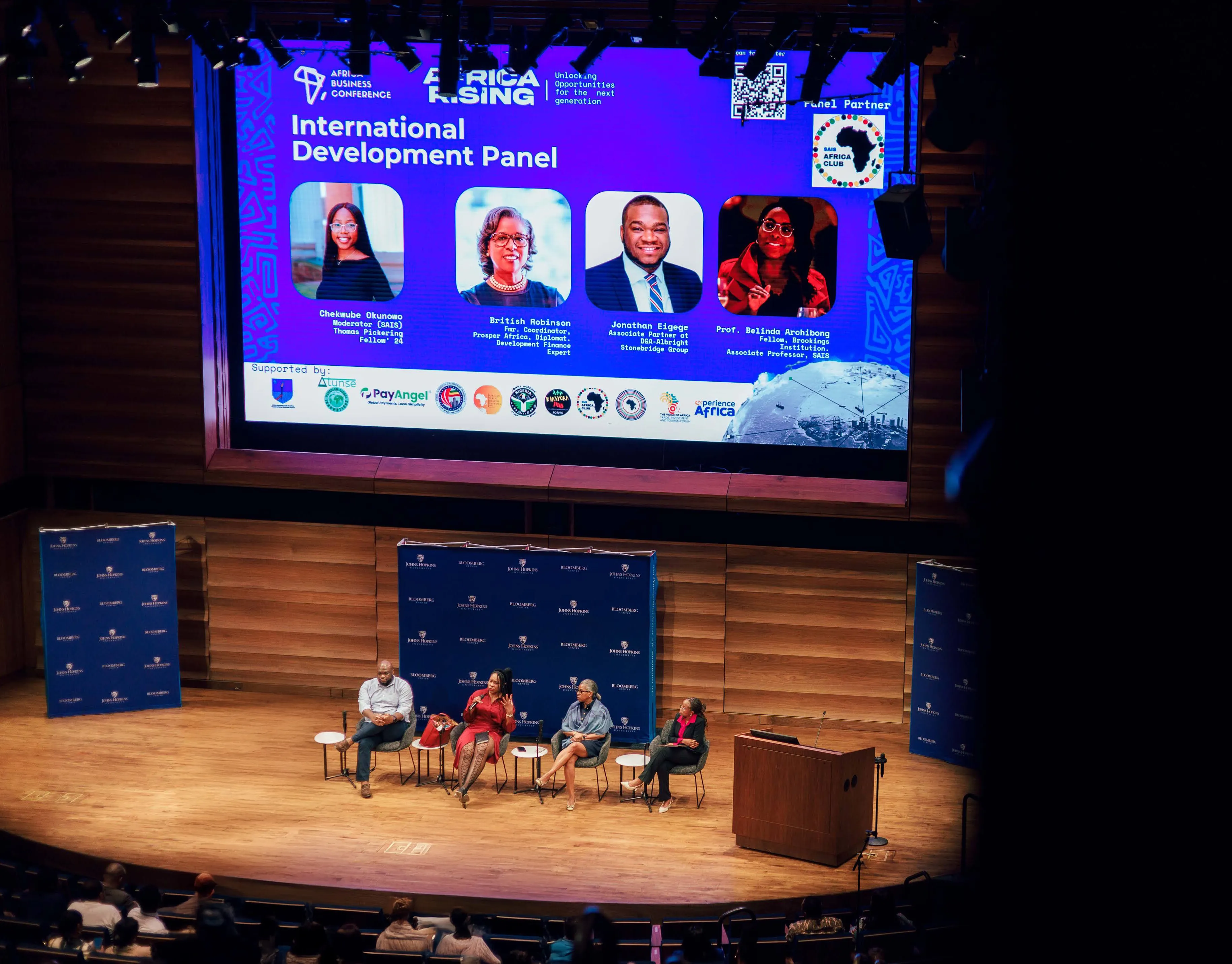 four people sitting in chairs on a stage. one person is holding and speaking into a microphone. a projector screen is in the background with their headshots