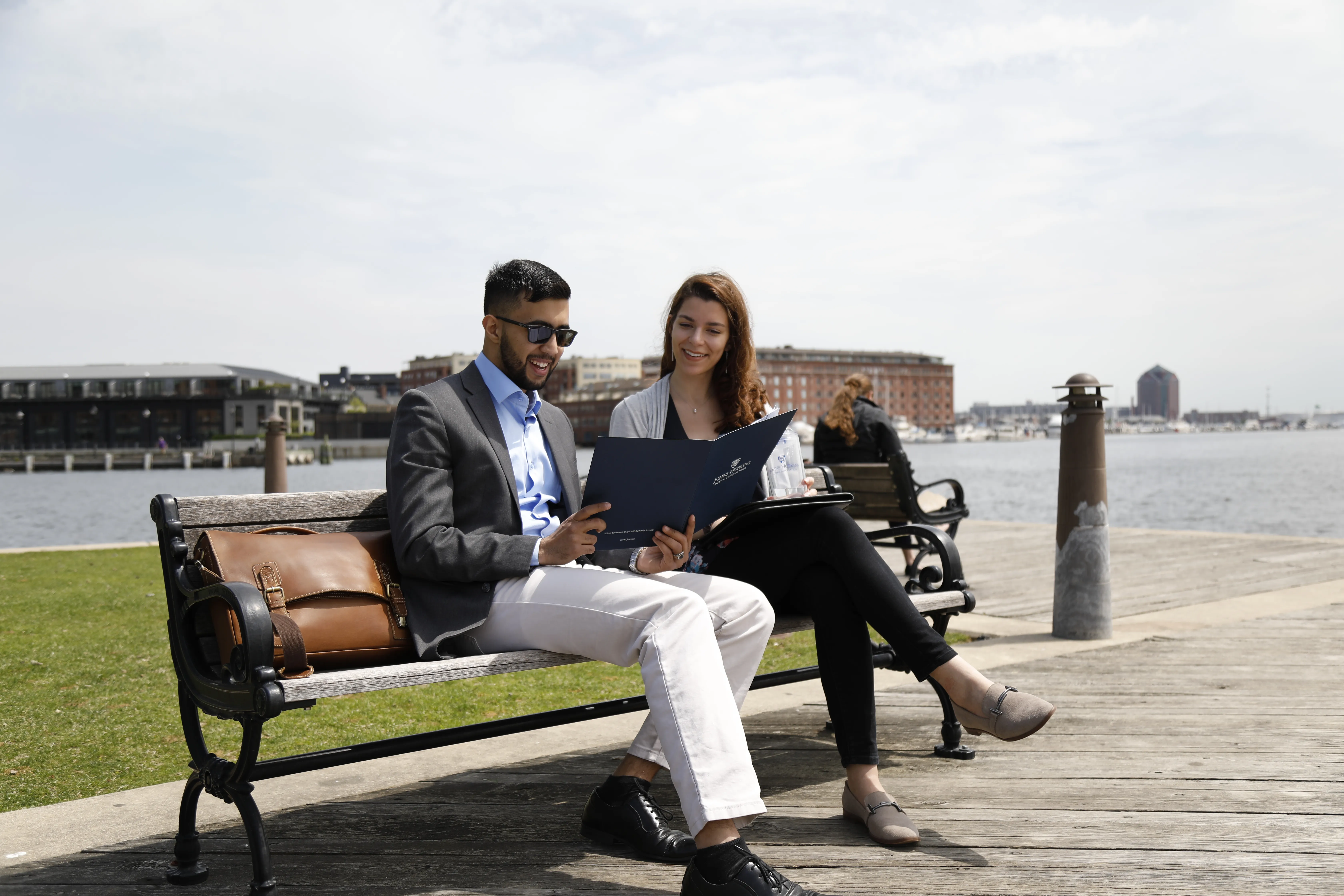 two people sitting on a bench outside in front of water, looking at a folder 