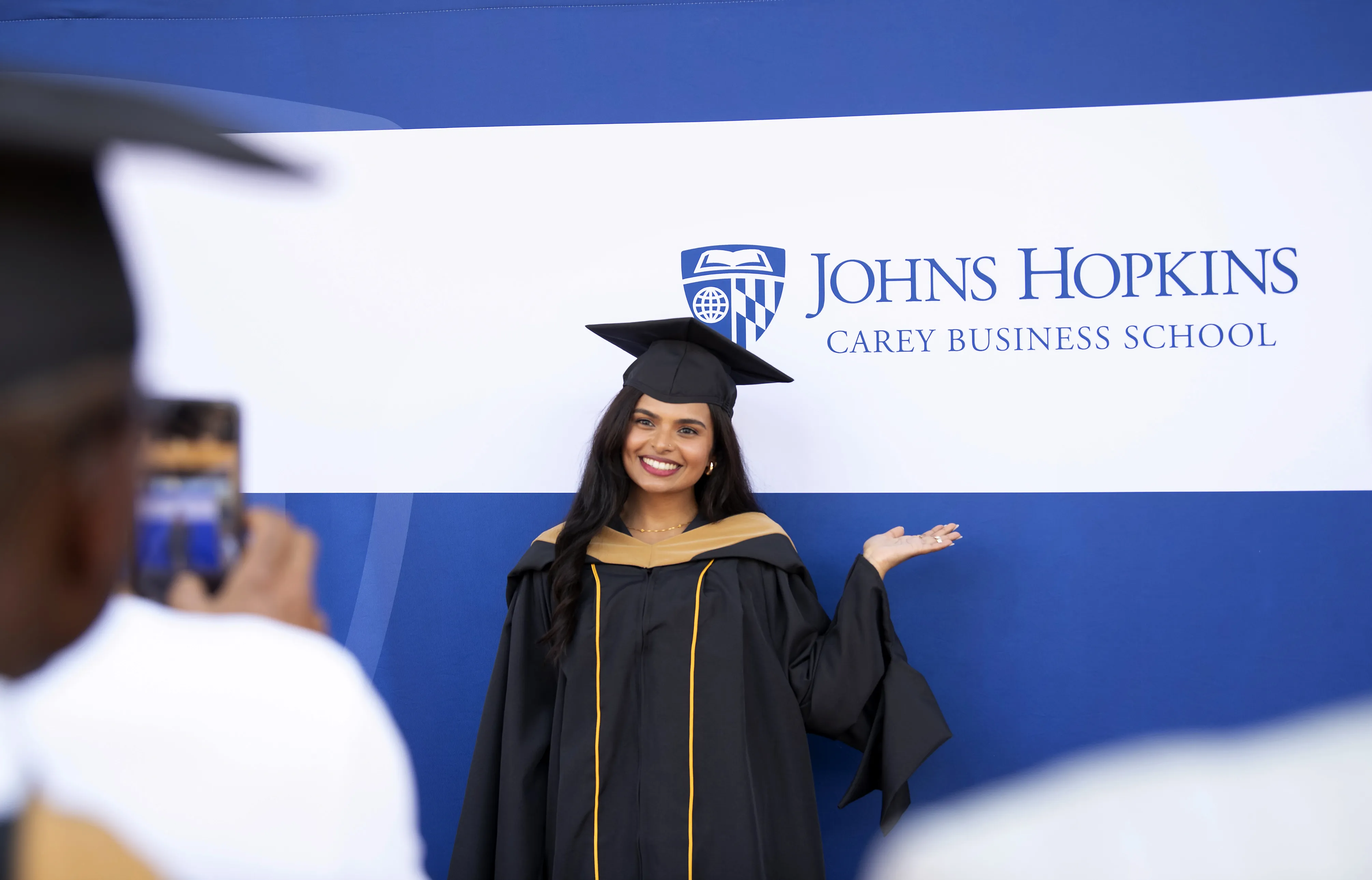person in graduation regalia psoing for a photo with a johns hopkins carey business school banner backdrop