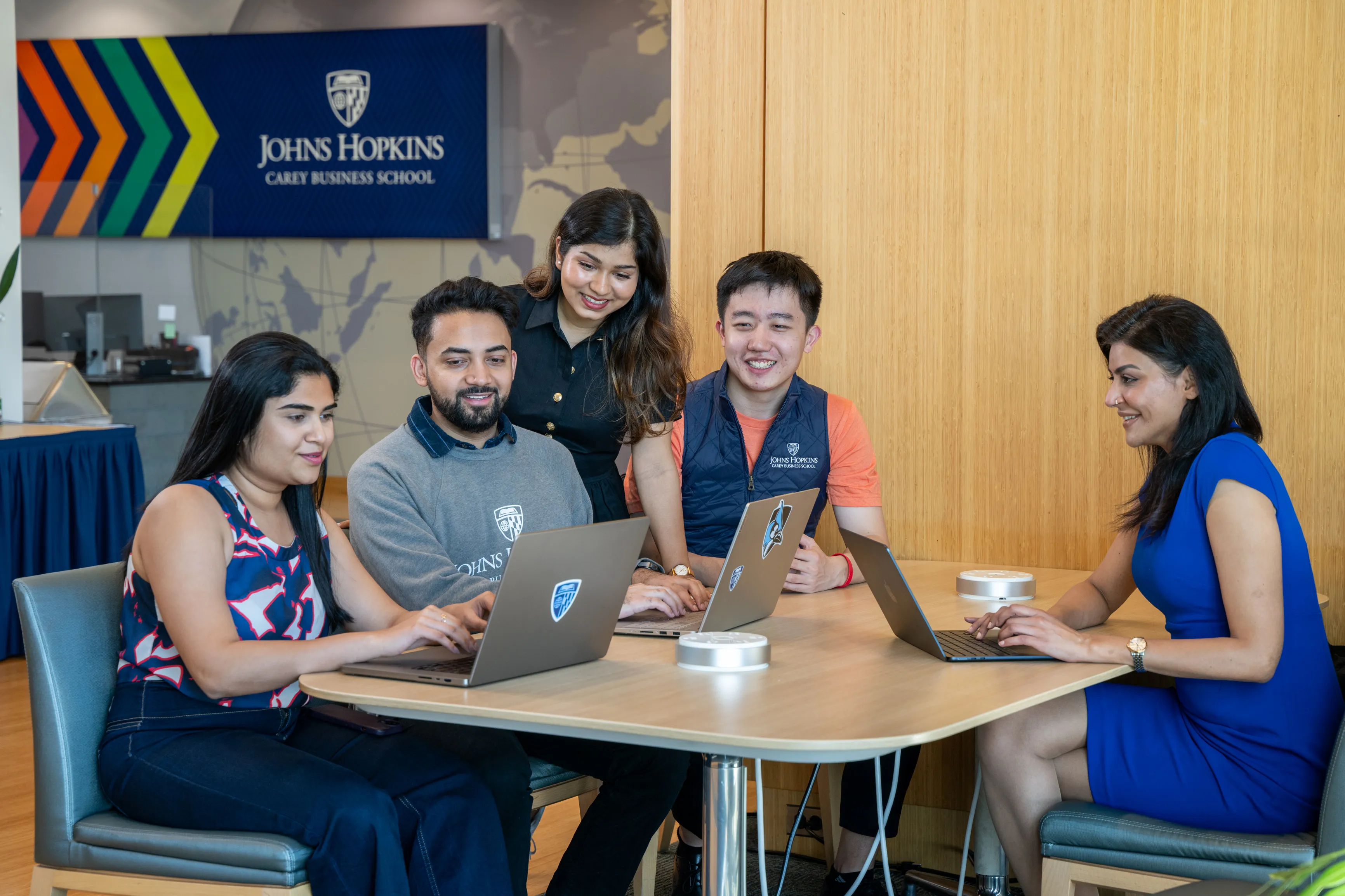 five people sitting around a desk with laptops out, talking to each other