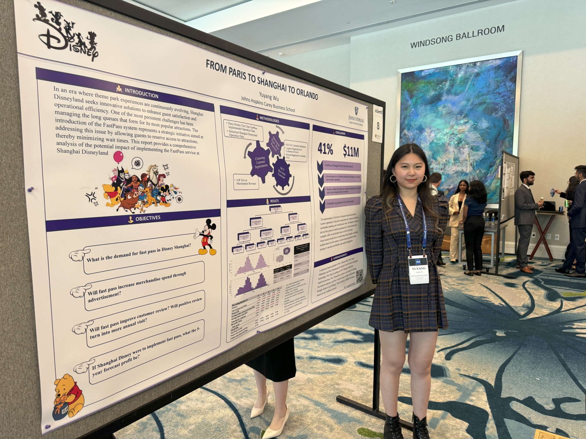 person standing in front of a large poster board smiling. people in the back also presenting posters