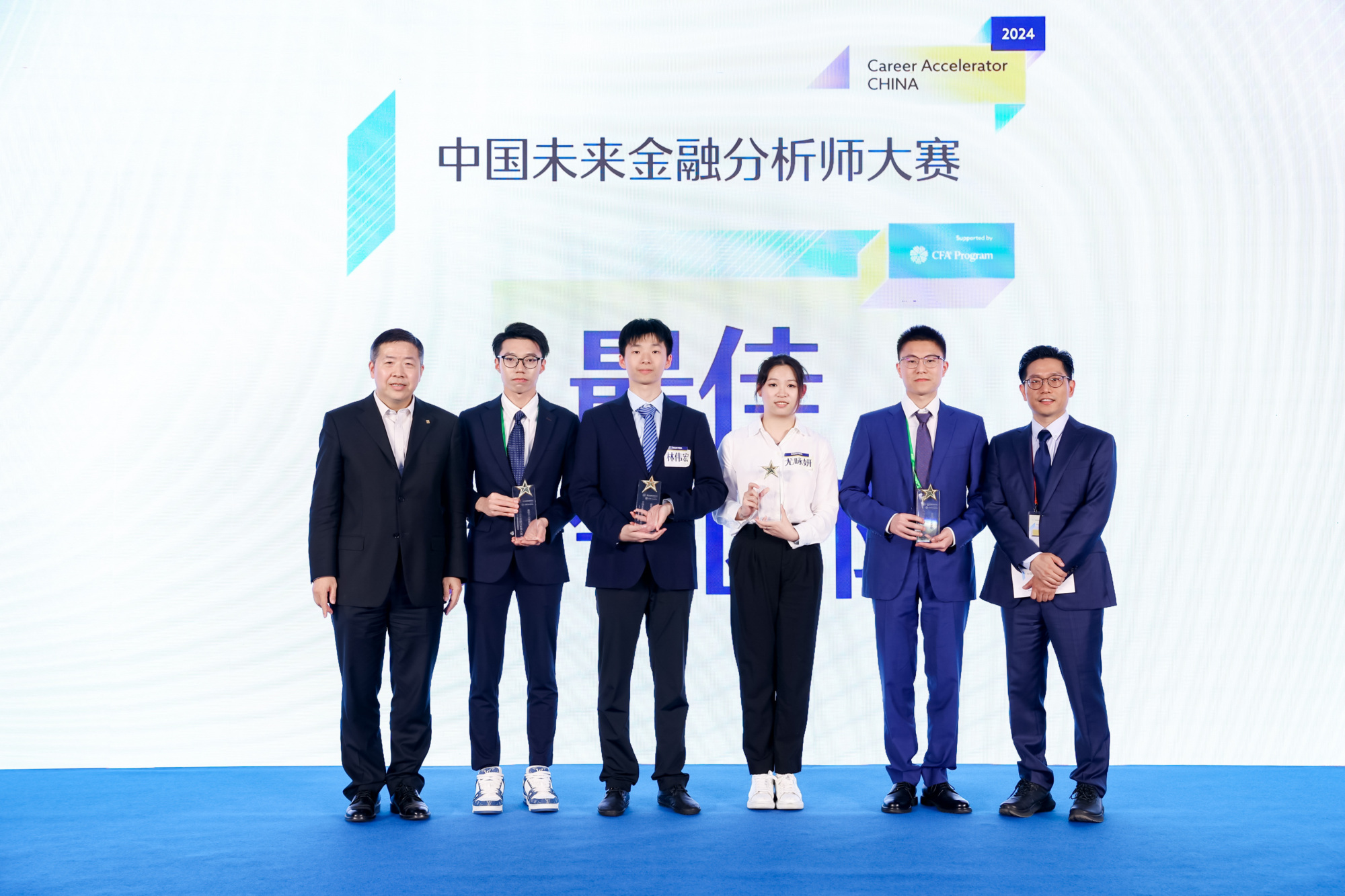 six people posing for a photo on a blue carpet with a large presentation on the screen behind them