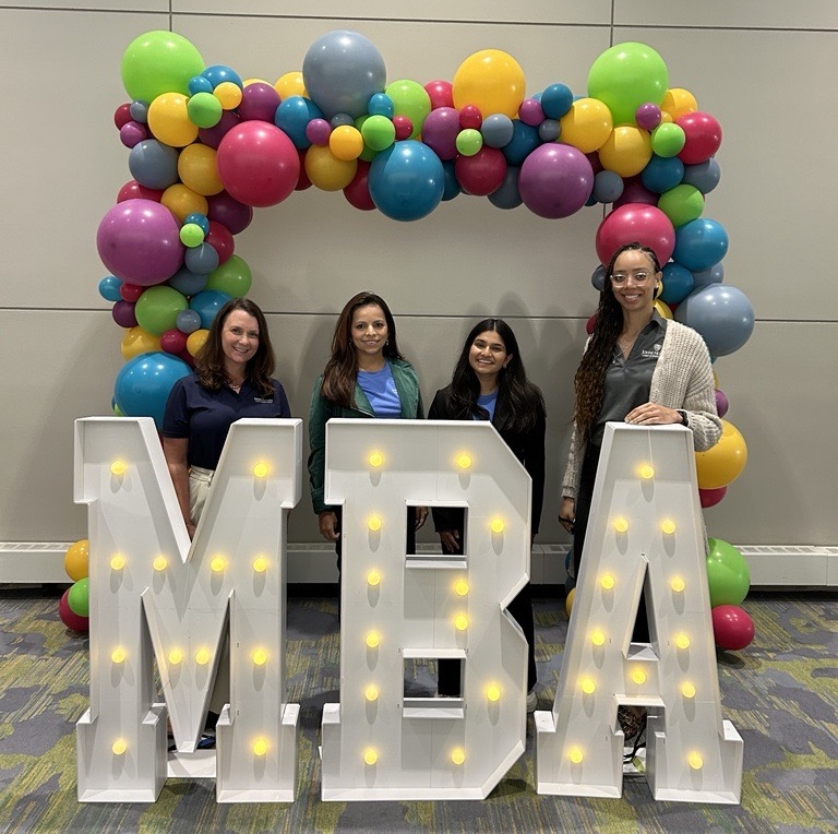 four people standing in back of big light up letters that spell "MBA" with a balloon arch in the back