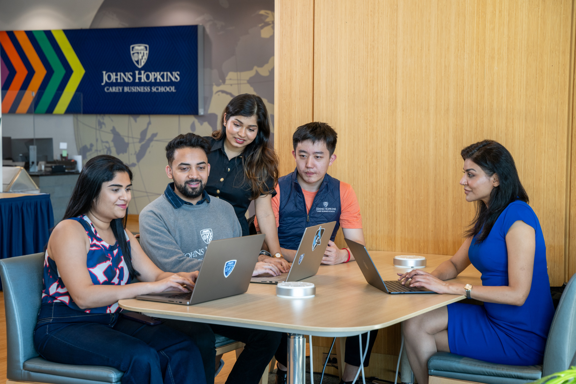 five people sitting at a table, four of them looking at a laptop and the other looking at their own laptop