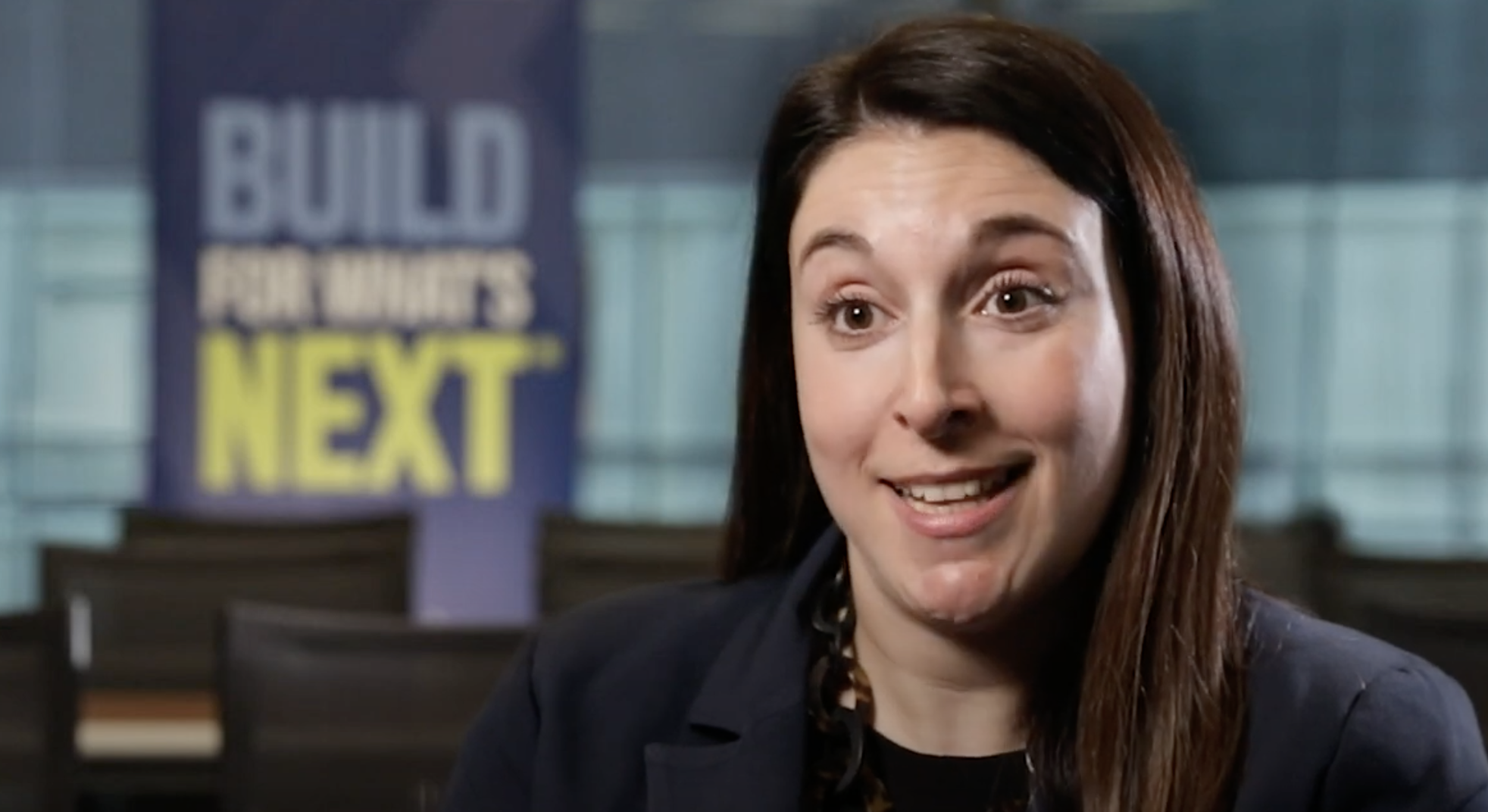 Person sitting in front of a build for what's next sign smiling while talking