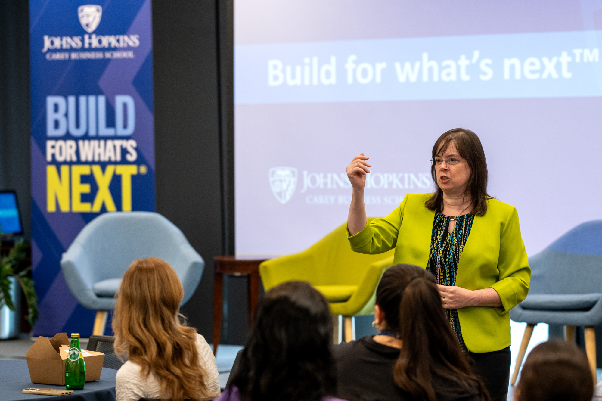 one person standing in front of a presentation screen speaking to an audience of people sitting at tables