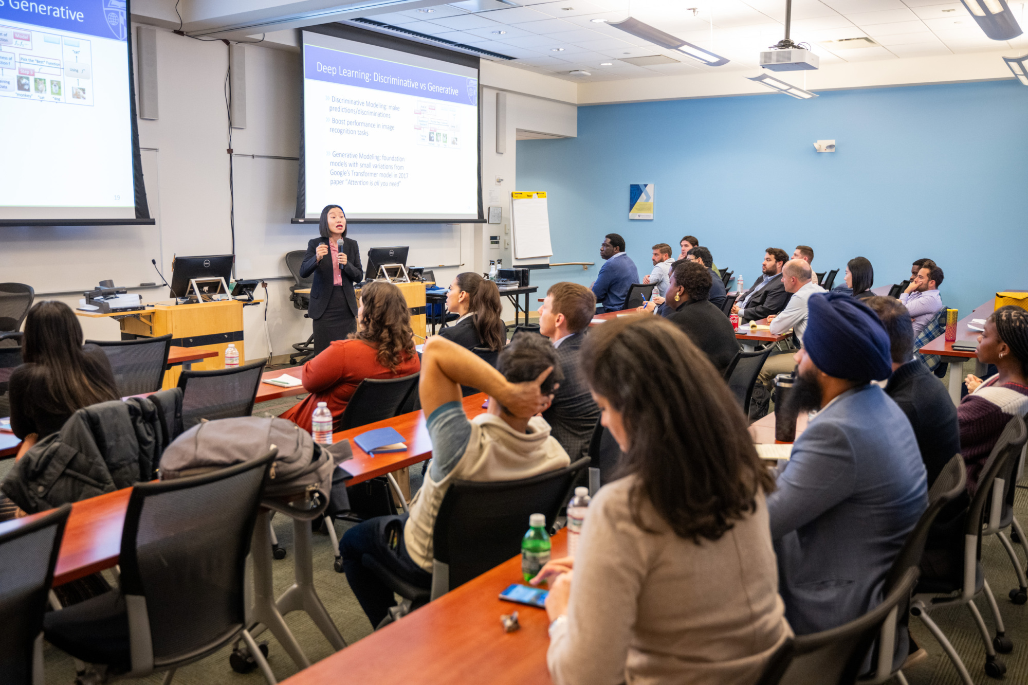 person holding microphone presenting to people sitting at tables in a classroom