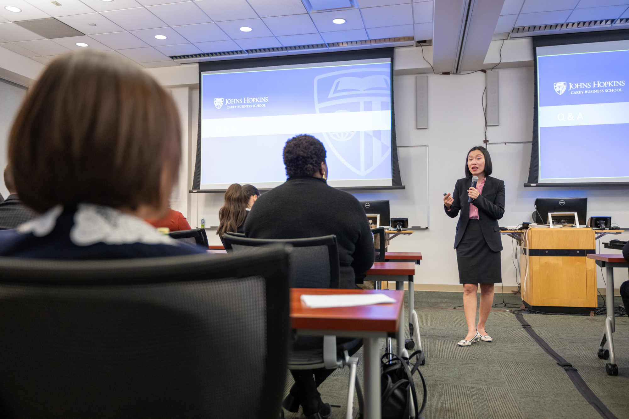 person standing with microphone speaking to people sitting at tables in a classroom. screen projectors in the background with presentation