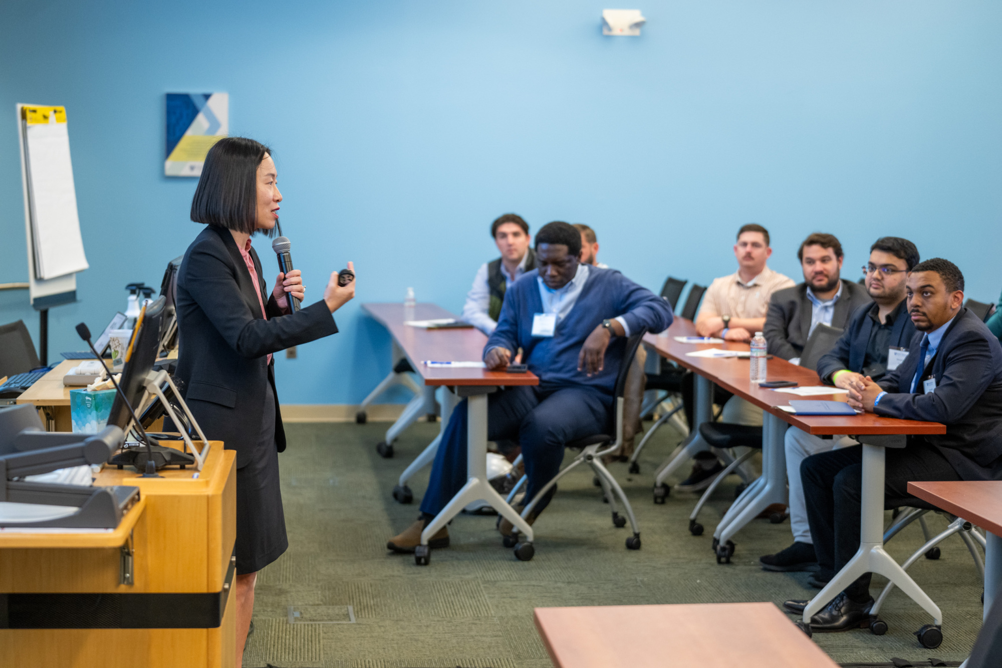 one person standing presenting with a microphone in hand to people sitting at desks in a classroom