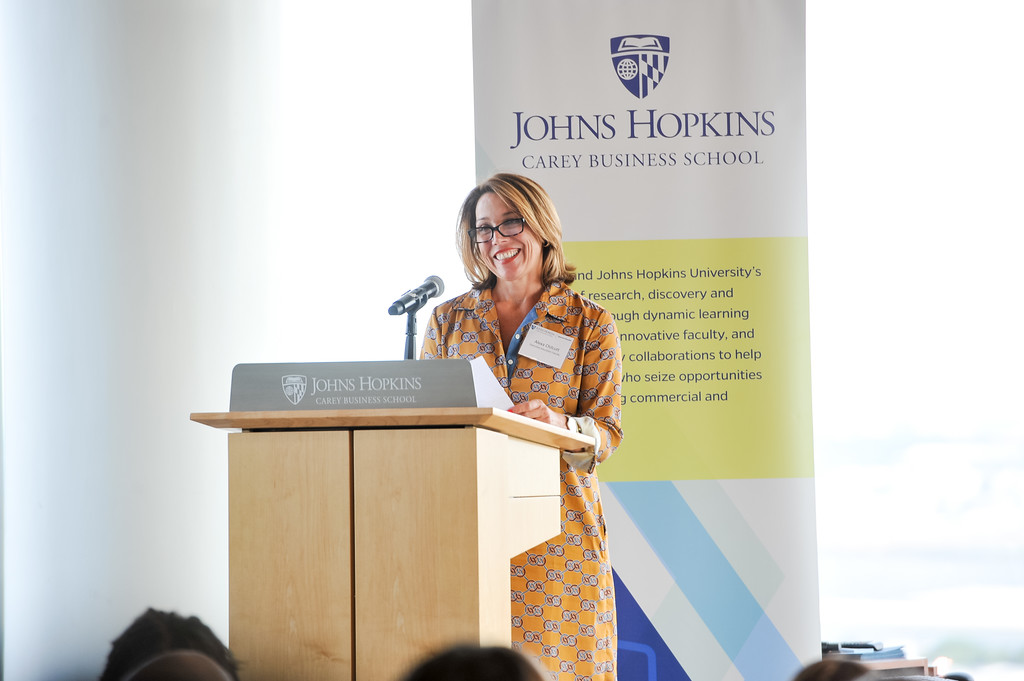 person standing a podium speaking into a microphone. Johns Hopkins Carey Business School logo in background