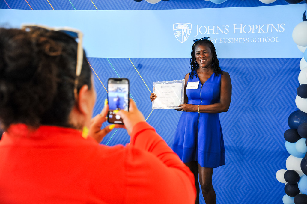 photo of a person talking a phone picture of someone standing in front of a Johns Hopkins Carey Business School banner holding a certificate