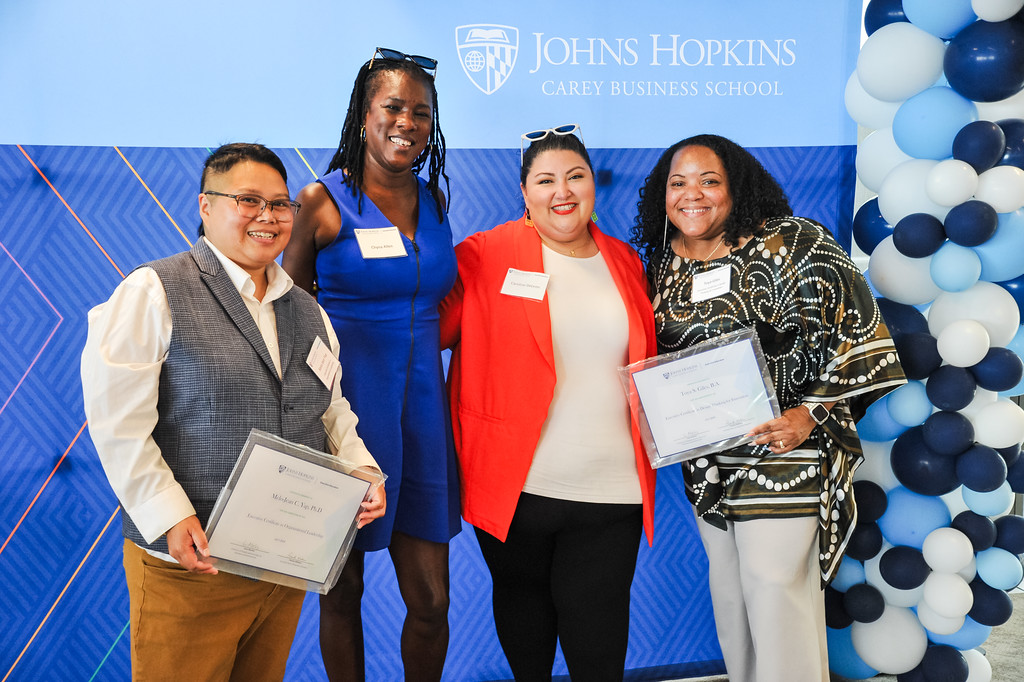 four people posing for photo, two are holding a certificate. Johns Hopkins Carey Business School banner behind them