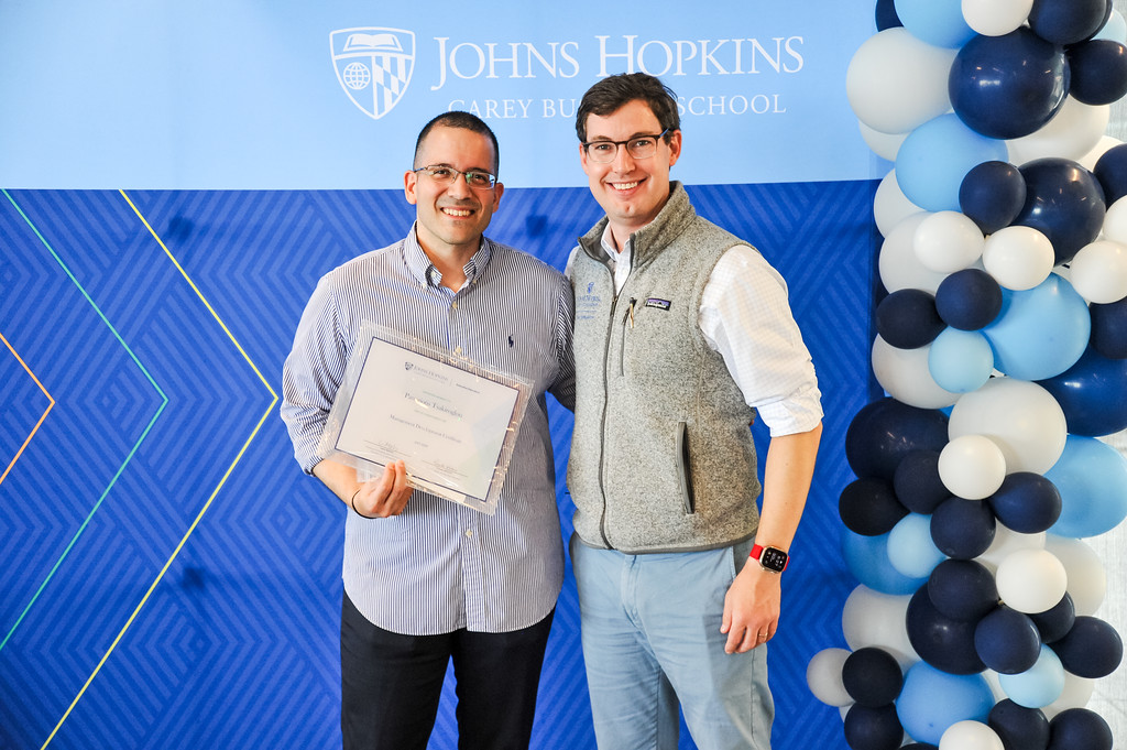 two people posing for photo, one is holding a certificate. Johns Hopkins Carey Business School banner behind them