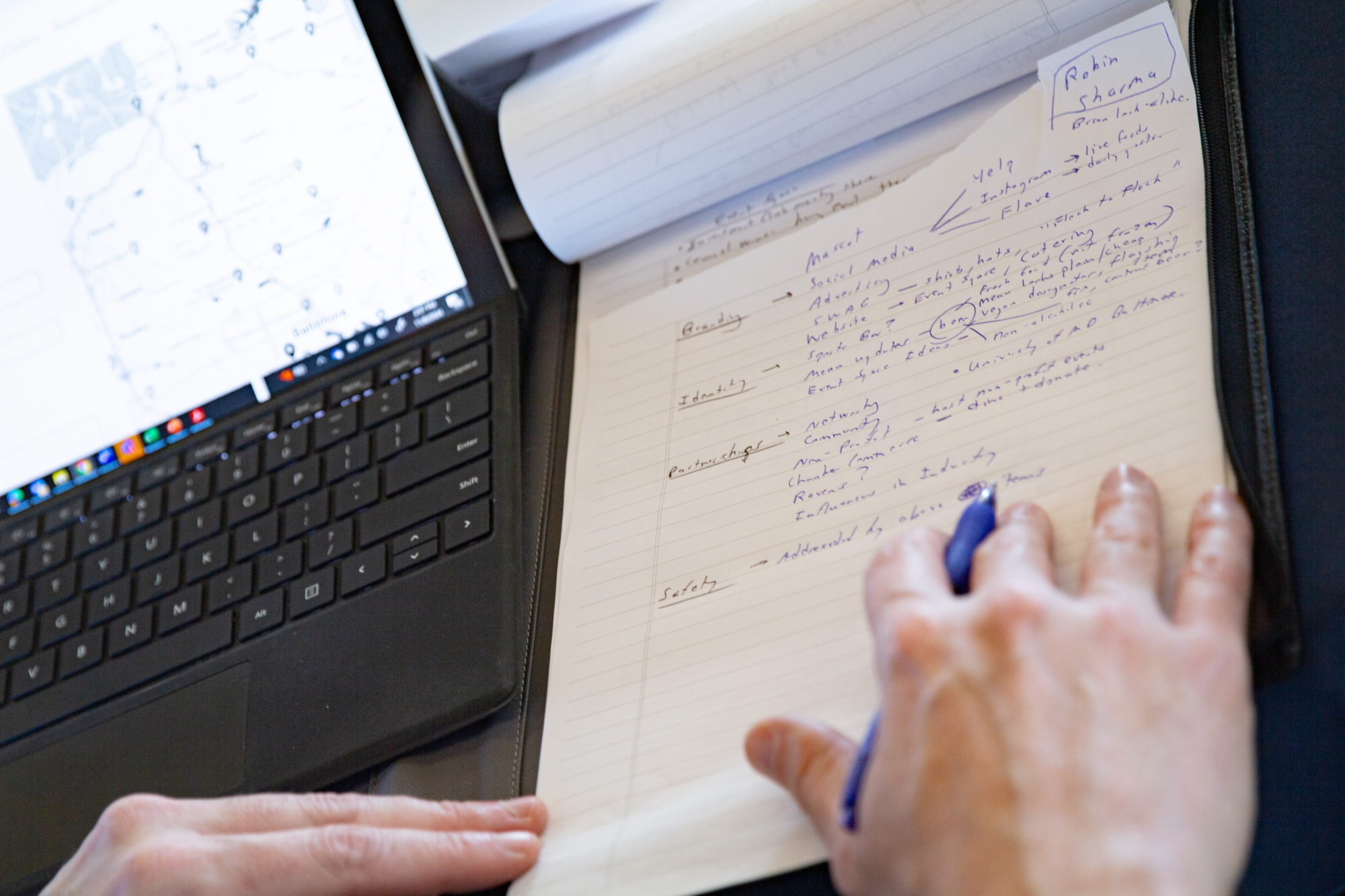 laptop and notepad next to each other zoomed in with a persons hand holding a pen on the notepad