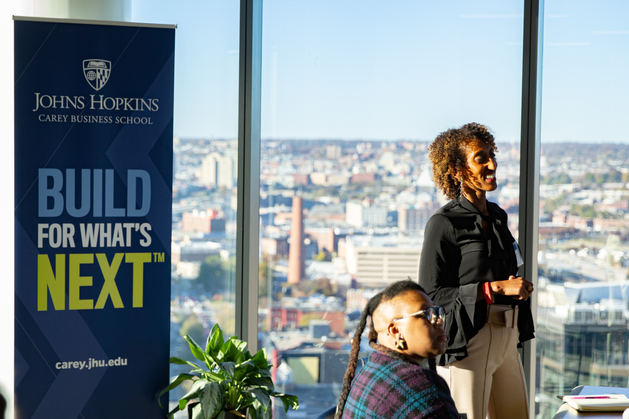 one person standing up, another sitting down. build for what's next banner in the background and the windows are glass with view of city